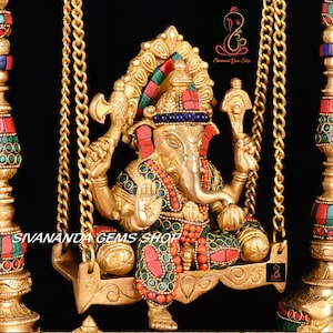 May include: A gold-colored Ganesha statue with red and green gemstones, sitting on a swing with gold chains. The statue is decorated with intricate details and is a traditional Hindu deity.