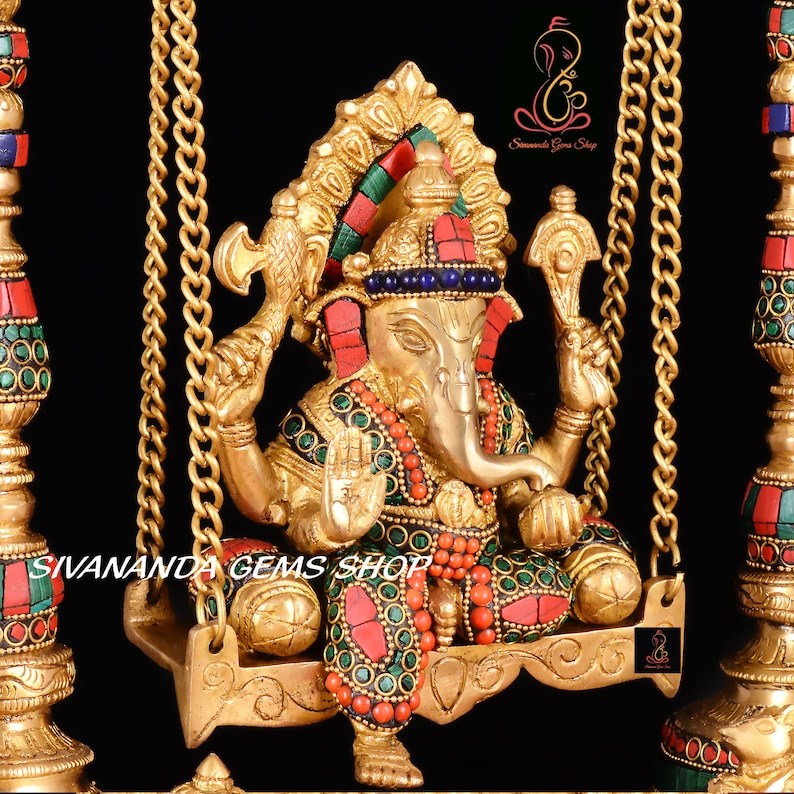May include: A gold-colored Ganesha statue with red and green gemstones, sitting on a swing with gold chains. The statue is decorated with intricate details and is a traditional Hindu deity.