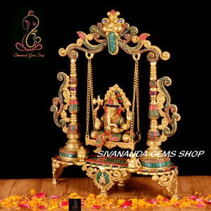 May include: Gold-toned metal sculpture of Ganesha, the Hindu god of wisdom and prosperity, sitting on a swing decorated with colorful gemstones. The swing is supported by two ornate pillars with intricate details. The sculpture is set against a black background.
