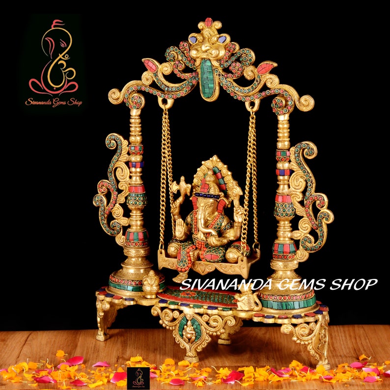 May include: Gold-toned metal sculpture of Ganesha, the Hindu god of wisdom and prosperity, sitting on a swing decorated with colorful gemstones. The swing is supported by two ornate pillars with intricate details. The sculpture is set against a black background.