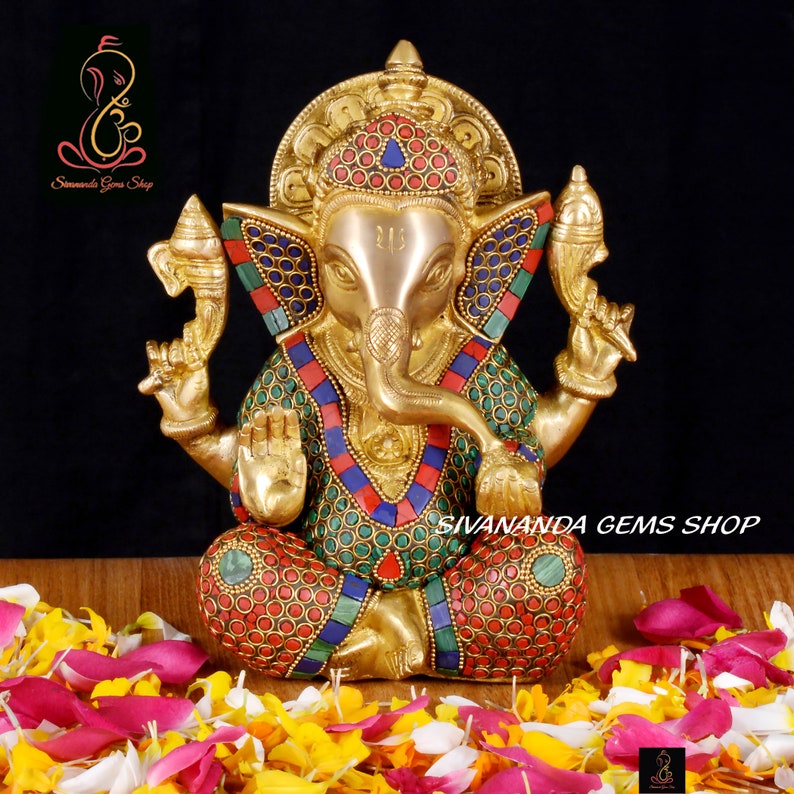 May include: A brass statue of the Hindu deity Ganesha, adorned with colorful inlaid stones. The statue is seated and features intricate details, including a large head and elephant trunk. The text "Sivananda Gems Shop" is visible.