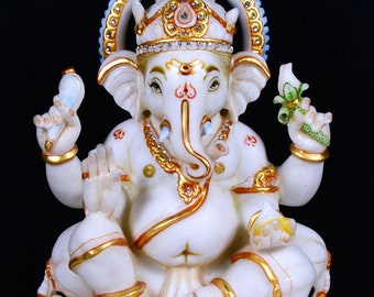 May include: White marble statue of Ganesha, the Hindu god of wisdom and prosperity, with a gold crown and a blue and gold halo. The statue is sitting on a white marble base.