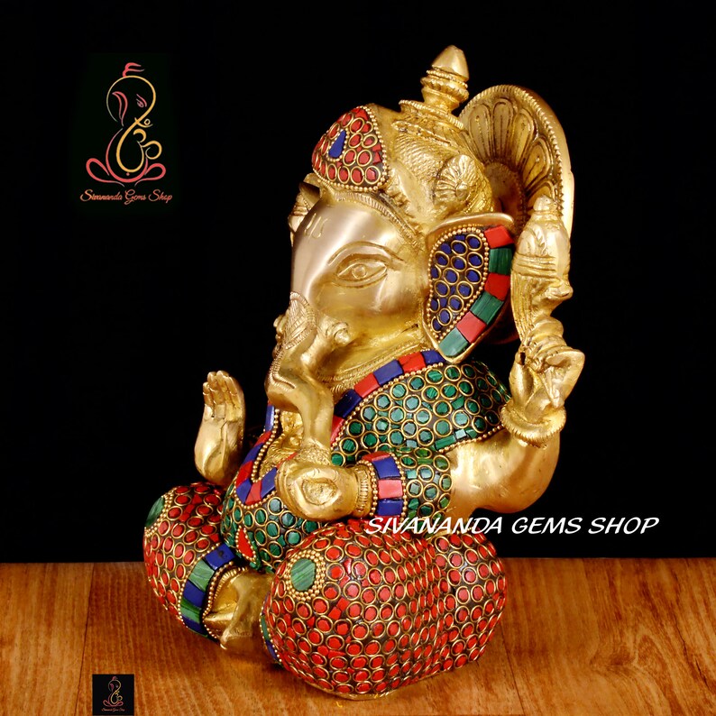 May include: A golden brass statue of the Hindu deity Ganesha, seated with intricate details. The statue features colorful inlaid stones in red, blue, and green. The text "Sivananda Gems Shop" is visible.