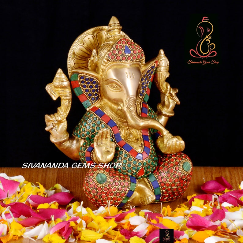 May include: A brass statue of the Hindu deity Ganesha, seated with intricate details. The statue is adorned with colorful inlaid stones and is surrounded by flower petals. The text "Sivananda Gems Shop" is visible.