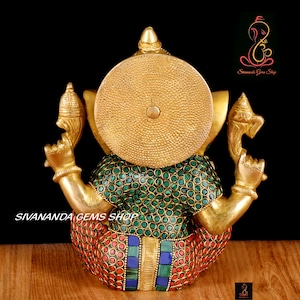 May include: A gold-colored statue of Ganesha, the Hindu god of wisdom and prosperity. The statue features a large, round head and multiple arms. The body is adorned with green, red, and blue gemstones. The text "SIVANANDA GEMS SHOP" is visible.