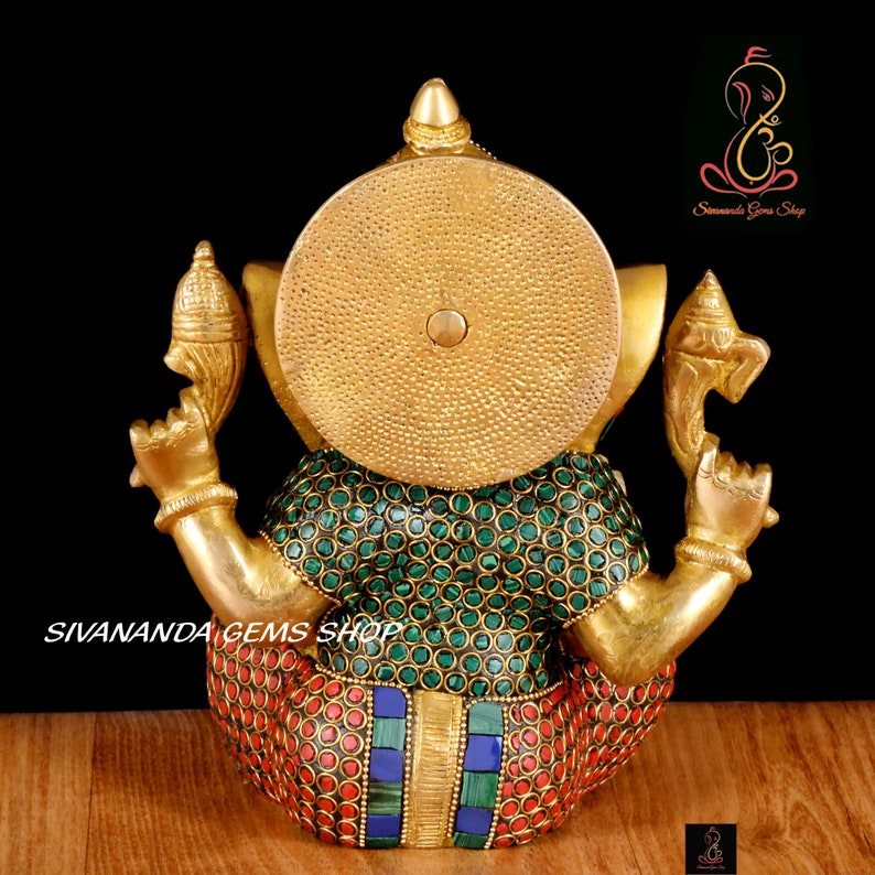 May include: A gold-colored statue of Ganesha, the Hindu god of wisdom and prosperity. The statue features a large, round head and multiple arms. The body is adorned with green, red, and blue gemstones. The text "SIVANANDA GEMS SHOP" is visible.