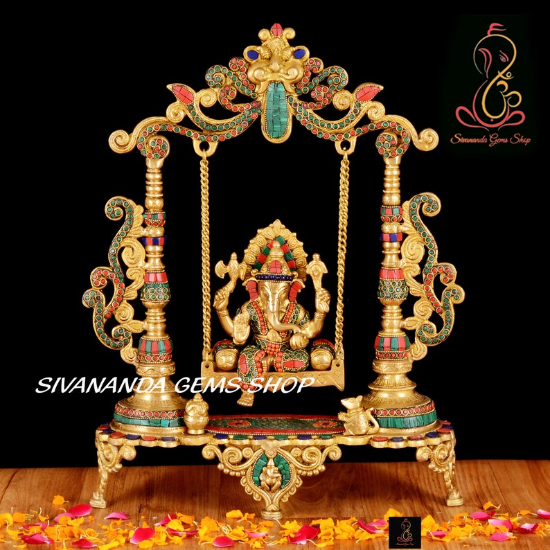 May include: Gold-toned metal sculpture of Ganesha, the Hindu god of wisdom and prosperity, sitting on a swing. The swing is decorated with colorful gemstones and intricate details. The sculpture is on a base with a small elephant figurine.