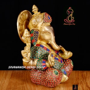 May include: A brass Ganesh statue with intricate details. The deity is adorned with colorful gemstones in red, green, and blue. The statue features a detailed crown and is seated in a meditative pose. The text "Sivananda Gems Shop" is visible.