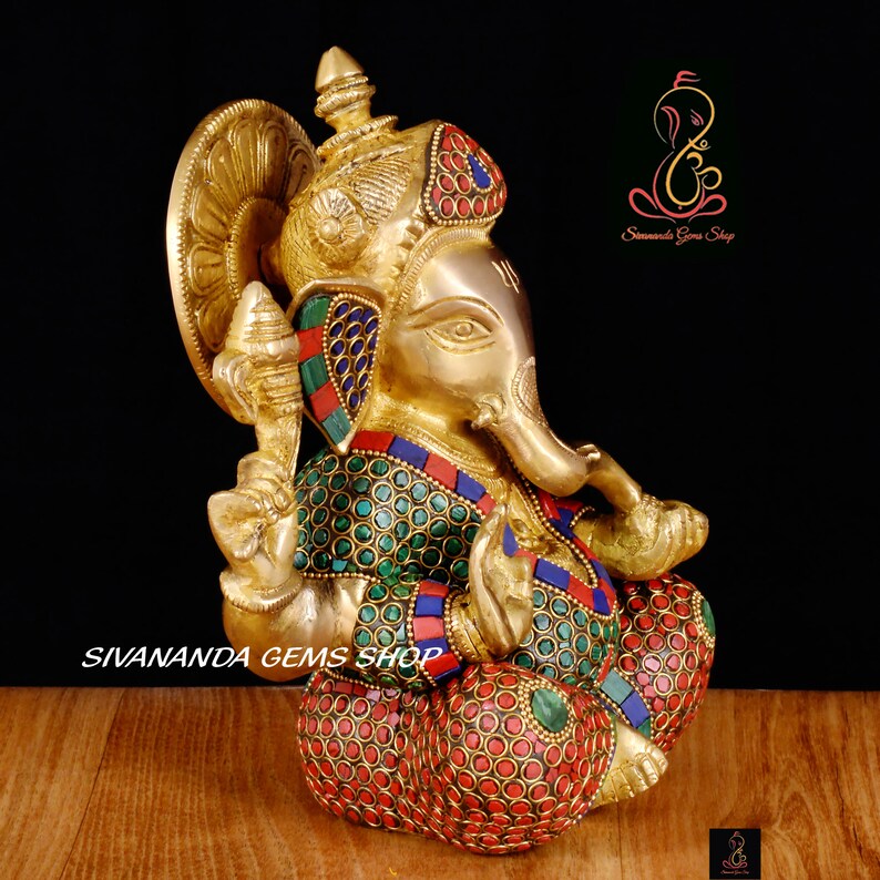 May include: A brass Ganesh statue with intricate details. The deity is adorned with colorful gemstones in red, green, and blue. The statue features a detailed crown and is seated in a meditative pose. The text "Sivananda Gems Shop" is visible.