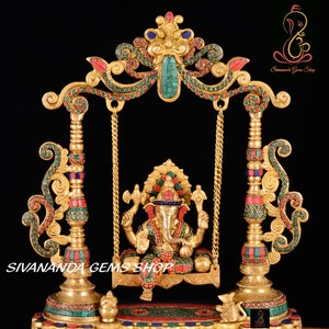 May include: A gold-colored metal statue of Ganesha, the Hindu god of beginnings, prosperity, and good fortune, sitting on a swing. The swing is decorated with red and green gemstones. The statue is on a base with a small elephant on each side. The text "Sivananda Gems Shop" is visible at the bottom of the image.