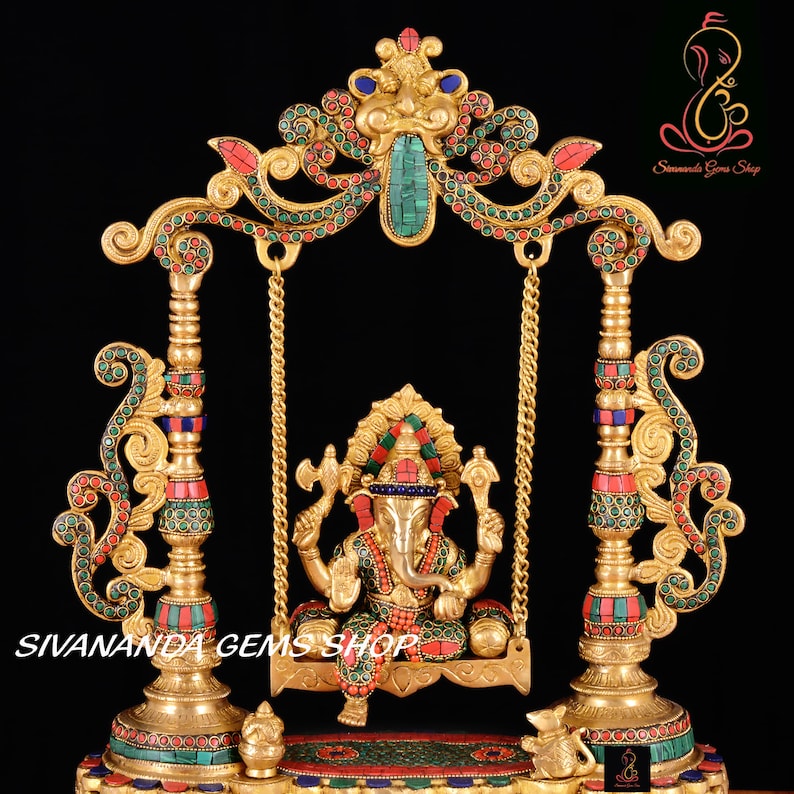 May include: A gold-colored metal statue of Ganesha, the Hindu god of beginnings, prosperity, and good fortune, sitting on a swing. The swing is decorated with red and green gemstones. The statue is on a base with a small elephant on each side. The text "Sivananda Gems Shop" is visible at the bottom of the image.