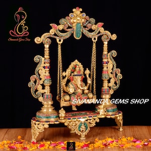 May include: A gold-colored metal sculpture of Ganesha, the Hindu god of wisdom and prosperity, sitting on a swing. The swing is decorated with colorful gemstones and intricate details. The sculpture is on a base with a similar design and is surrounded by pink and orange flower petals.