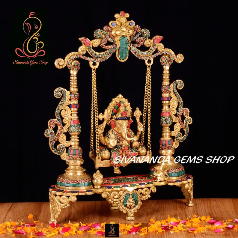 May include: A gold-colored metal sculpture of Ganesha, the Hindu god of wisdom and prosperity, sitting on a swing. The swing is decorated with colorful gemstones and intricate details. The sculpture is on a base with a similar design and is surrounded by pink and orange flower petals.