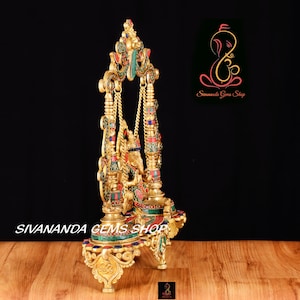 May include: A gold-colored metal statue of Ganesha, the Hindu god of wisdom and prosperity, sitting on a swing. The statue is decorated with colorful gemstones and intricate details. The statue is on a wooden surface.