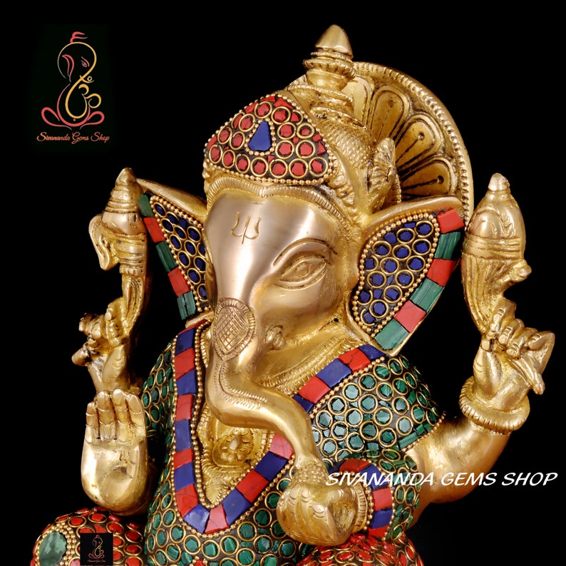 May include: A detailed brass statue of the Hindu deity Ganesha, adorned with colorful gemstones. The statue features a gold finish with intricate detailing, including a crown and decorative elements. The text "Sivananda Gems Shop" is visible.