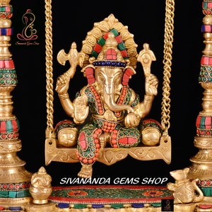 May include: A gold-colored statue of Ganesha, the Hindu god of wisdom and prosperity, sitting on a swing. The statue is decorated with colorful gemstones and intricate details. The swing is made of gold chains and has a decorative base.