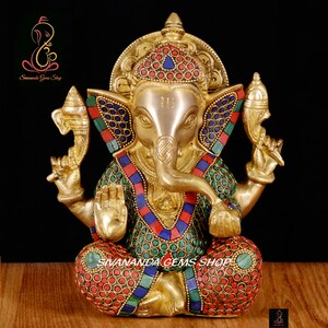 May include: A gold-colored statue of Ganesha, the Hindu god of wisdom, with intricate details. The statue features colorful inlaid stones in red, blue, and green. The text "Sivananda Gems Shop" is visible.