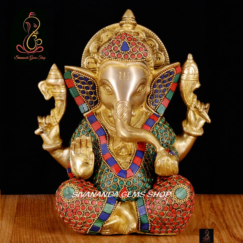 May include: A gold-colored statue of Ganesha, the Hindu god of wisdom, with intricate details. The statue features colorful inlaid stones in red, blue, and green. The text "Sivananda Gems Shop" is visible.