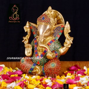 May include: A brass statue of the Hindu deity Ganesha, adorned with colorful inlaid stones. The statue is seated and surrounded by flower petals. The text "Sivananda Gems Shop" is visible.
