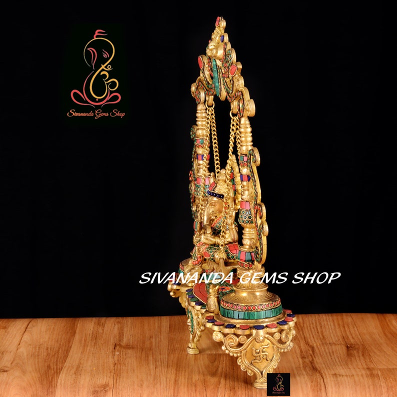 May include: A gold-colored metal statue of Ganesha, the Hindu god of wisdom and prosperity, sitting on a swing. The statue is decorated with colorful gemstones and is on a base with three legs. The statue is on a wooden surface.  Sivananda Gems Shop is printed on the base.