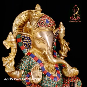 May include: A golden brass statue of the Hindu deity Ganesha, adorned with colorful inlaid gemstones. The statue features intricate details, including a large elephant head, multiple arms, and a seated posture. The text "SIVANANDA GEMS SHOP" is visible at the bottom.