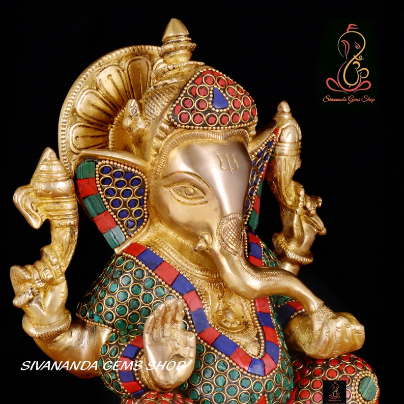 May include: A golden brass statue of the Hindu deity Ganesha, adorned with colorful inlaid gemstones. The statue features intricate details, including a large elephant head, multiple arms, and a seated posture. The text "SIVANANDA GEMS SHOP" is visible at the bottom.