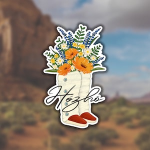 May include: White cowboy boots with a bouquet of blue, yellow, and white flowers inside. The boots have a red sole and the word "Hózhó" is written in black cursive on the side.