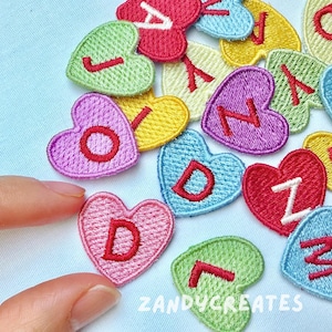 May include: Embroidered heart-shaped patches in various colors, each with a letter in a contrasting color. The patches are arranged on a light blue surface. The text "ZANDYCREATES" is visible.