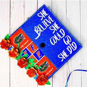 May include: Blue graduation cap decorated with glitter, white text that reads "She believed she could, she did", and a Florida Gators logo. The cap is adorned with orange paper flowers.