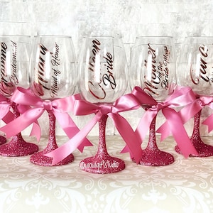 May include: Set of six pink glitter wine glasses with pink ribbon bows. Each glass has a gold vinyl decal with a different title, including "Bride", "Maid of Honor", and "Bridesmaid".