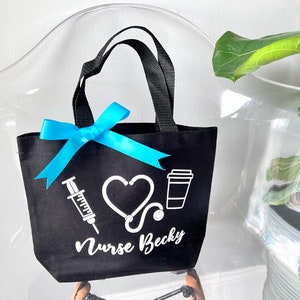 May include: Black tote bag with white text and graphics. The text reads "Nurse Becky" and the graphics include a heart, a stethoscope, a syringe, and a coffee cup.
