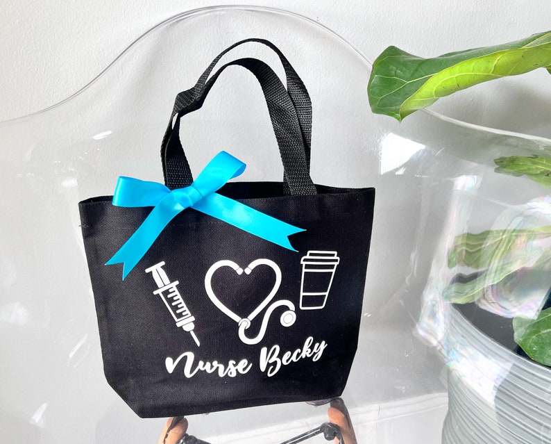 May include: Black tote bag with white text and graphics. The text reads "Nurse Becky" and the graphics include a heart, a stethoscope, a syringe, and a coffee cup.