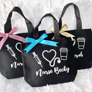May include: Three black tote bags with white designs. The bags have black handles and are decorated with a pink, blue, and gold ribbon. The designs include a heart, a stethoscope, a syringe, and a coffee cup. The text on the bags reads "Nurse Becky" and "Nurse".