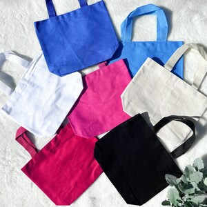 May include: Seven tote bags in various colours, including white, black, pink, blue, and light blue. The bags are made of canvas and have a simple design.