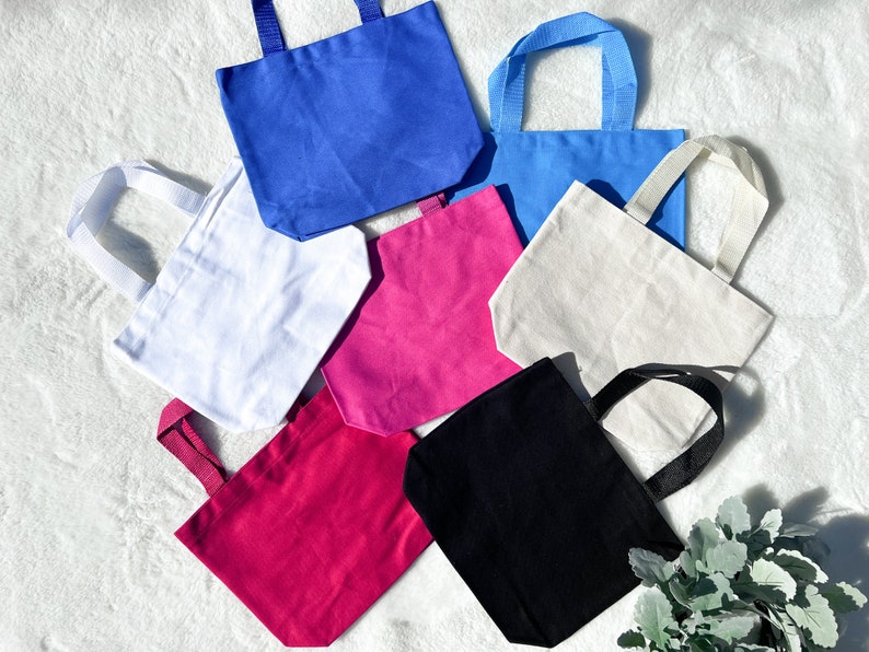 May include: Seven tote bags in various colours, including white, black, pink, blue, and light blue. The bags are made of canvas and have a simple design.