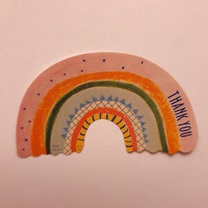 Op de afbeelding: Een kleurrijke kaart in de vorm van een regenboog met de tekst "THANK YOU" gedrukt aan de rechterkant. De regenboog heeft een geel smiley-gezicht aan de onderkant.