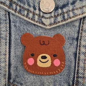 Può includere: Una toppa a forma di orso marrone con guance rosa e un viso sorridente è cucita su una tasca di giacca di jeans. La giacca ha un bottone argentato con "Denim Co."