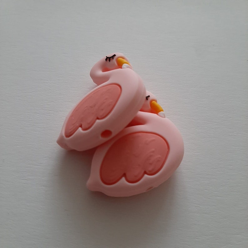 May include: Two pink silicone flamingo toys with yellow beaks and black eyes. The toys are stacked on top of each other.