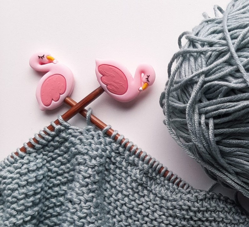 May include: Two pink flamingo knitting needles with a gray knitted fabric in the background. The needles are crossed and the fabric is a close-up of the knit stitches.