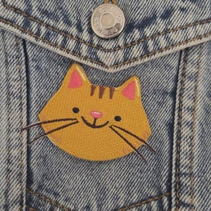 Peut inclure: Gros plan d'une poche de veste en jean avec un écusson de chat jaune. L'écusson représente un visage de chat souriant avec des oreilles et un nez roses, et des moustaches noires. La veste a un bouton argenté en haut de la poche.