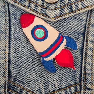Puede incluir: Parche bordado de cohete con detalles rojos, azules y blancos. El parche está adherido a una chaqueta vaquera. El cohete tiene una llama roja y una ventana circular verde.