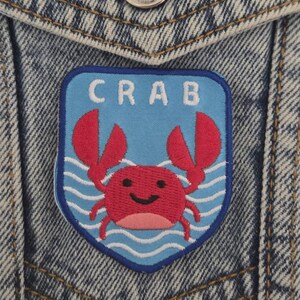 Op de afbeelding: Een blauw en rode krab patch met het woord "CRAB" in witte letters. De patch heeft een cartoon krab met rode klauwen en een lachend gezicht, tegen een lichtblauwe achtergrond met witte golfdetails. De patch is op een spijkerjas genaaid.