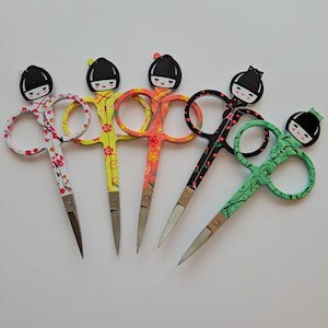 May include: Five pairs of colourful, decorative scissors with a Japanese doll design. Each pair has a different colour and pattern, including yellow, orange, green, and black.