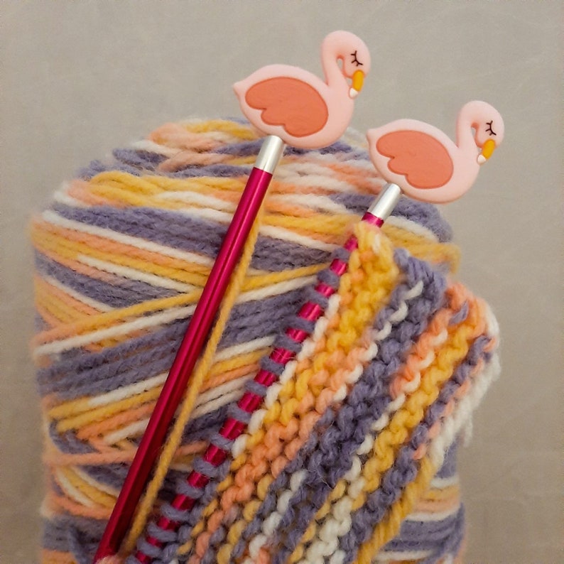 May include: A ball of multi-colored yarn with two pink flamingo knitting needles. The yarn is a mix of white, purple, orange, and yellow.