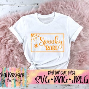 以下が含まれることがあります： 「Spooky Babe」とオレンジ色のグラフィックが書かれた白いTシャツ。コーナーにはクモの巣とクモがあります。