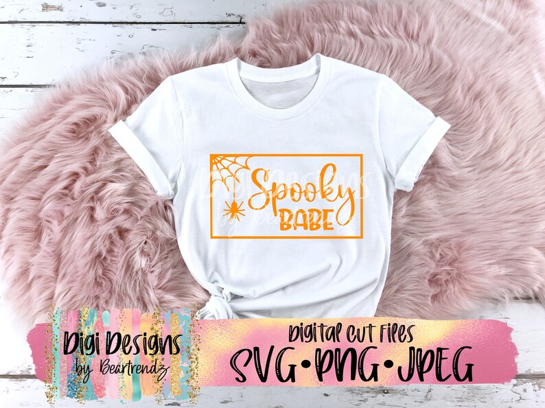 以下が含まれることがあります： 「Spooky Babe」とオレンジ色のグラフィックが書かれた白いTシャツ。コーナーにはクモの巣とクモがあります。