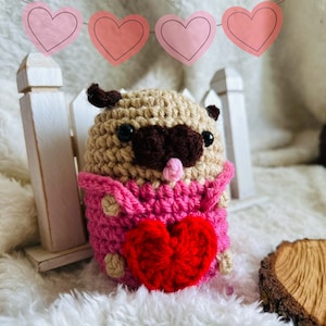 May include: A crocheted pug wearing a pink sweater and holding a red heart. The pug is sitting in front of a white picket fence with a pink heart garland.