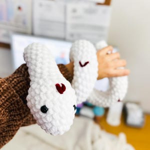 May include: A white crocheted snake with red hearts on its body. The snake is being held by a person's hand.