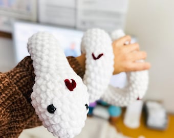 May include: A white crocheted snake with red hearts on its body. The snake is being held by a person's hand.