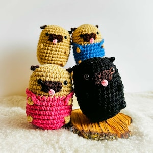 May include: Four crocheted pug dog plushies in different colors. The pugs are wearing different colored outfits. The pugs are sitting on a wooden surface.